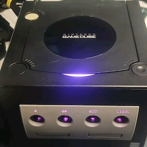 May include: A black Nintendo GameCube console with the text "NINTENDO GAMECUBE" on the top. The console has a silver faceplate with four controller ports and a disc drive slot. The power button glows purple.