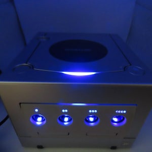 Może przedstawiać: Srebrna konsola do gier wideo z okrągłą tacą na dyski na górze i czterema świecącymi niebieskimi światłami z przodu. Konsola jest podświetlona niebieskim światłem, tworząc futurystyczną estetykę.