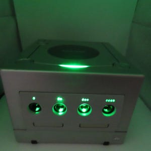 May include: A silver-colored video game console with a green glowing power button and controller ports. The console has a circular disc tray on top and four controller ports on the front, all illuminated with green lights.