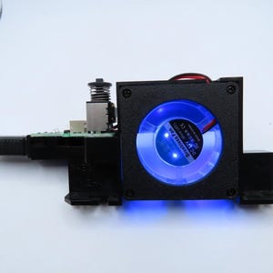 May include: A black electronic device with a blue LED fan. The device has a square housing with a circular fan visible through the front. A circuit board and a spring are also visible. A black cable extends from the device.