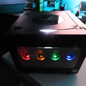 May include: A black video game console with a silver top and four illuminated buttons in red, orange, green, and blue. The console has a sleek, modern design with a circular disc tray and a power indicator light.