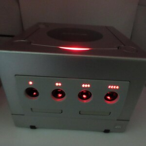 Può includere: Console per videogiochi argentata con una luce rossa e quattro spie rosse. La console ha un vano disco e un pulsante di accensione. La console ha una forma a cubo.