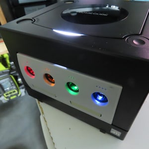 May include: A black GameCube console with a silver faceplate. The faceplate has four colored lights: red, orange, green, and blue. The console has a disc tray and a power button. The top of the console has the words "GameCube" printed on it.