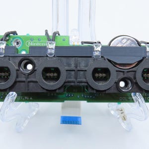 May include: A green circuit board with black ports and the Nintendo logo. The board has a battery and various electronic components. The board is supported by clear plastic stands.
