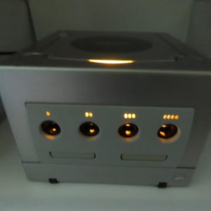 May include: Silver video game console with a black power cord. The console has a rectangular shape with a central panel featuring four circular buttons and indicator lights. The top has a circular disc tray.