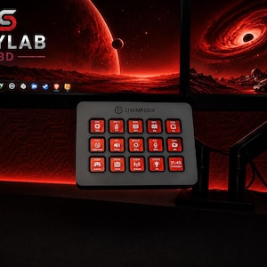 Può includere: Un controller Stream Deck bianco con quindici pulsanti quadrati con icone e testo retroilluminati in rosso. Il dispositivo è montato su un braccio regolabile, posizionato sotto due monitor di computer con uno sfondo spaziale rosso e il testo SKYLAB 3D.