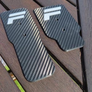 May include: Two carbon fiber automotive parts with a black and silver weave pattern. Each part has a white "IL" logo and a hole for mounting. The parts are displayed on a wooden surface.
