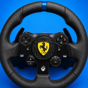 Könnte beinhalten: Ein schwarzes Rennlenkrad mit strukturiertem Ledergriff, einer blauen Markierung oben in der Mitte und einem gelben Ferrari-Logo in der Mitte. Das Gerät verfügt über eine Xbox-Taste, farbige A-, B-, X-, Y-Tasten und verschiedene Steuerelemente.