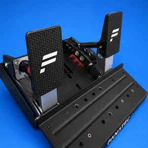 Op de afbeelding: Een zwarte metalen racepedaalset met twee pedalen voorzien van getextureerde zeshoekige oppervlakken en witte logo's. De basisplaat bevat de tekst FANATEC en Skylab3D, met meerdere ronde montagegaten en zichtbare mechanische veren.