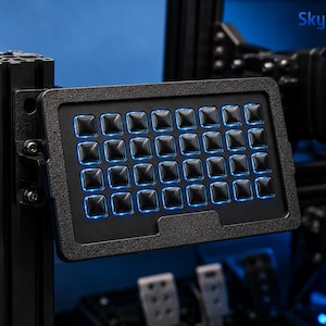 Puede incluir: Una caja de botones rectangular negra con una cuadrícula de treinta y dos botones cuadrados, cada uno con retroiluminación LED azul. El dispositivo está montado en un marco de aluminio negro y muestra el texto Skylab3D en la esquina superior derecha.