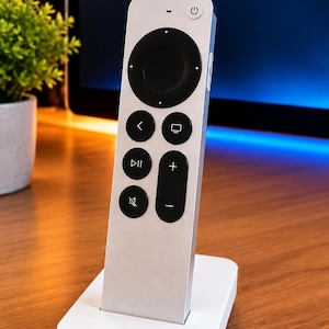 Può includere: Un telecomando argentato con un pad di navigazione circolare nero e pulsanti neri per accensione, indietro, home, riproduzione-pausa, volume e muto, posizionato verticalmente su una base di ricarica bianca rettangolare.