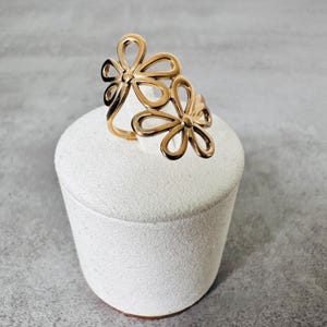 Peut inclure: Une bague dorée avec un motif floral, comprenant deux motifs de fleurs ajourées. La bague est présentée sur un socle cylindrique blanc sur fond gris. Les fleurs ont des pétales incurvés et une apparence délicate.