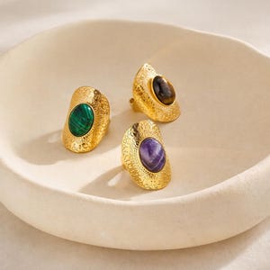 Peut inclure: Trois bagues dorées avec des pierres précieuses ovales. L'une des bagues présente une pierre verte, une autre une pierre brune et la troisième une pierre violette. Chaque bague a une bande texturée et à motifs.