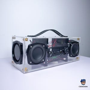 May include: A rectangular portable speaker with a transparent acrylic housing, featuring two black circular speakers and a central control panel with a Bluetooth icon, volume knob, and various input ports. A black carrying handle is attached to the top.