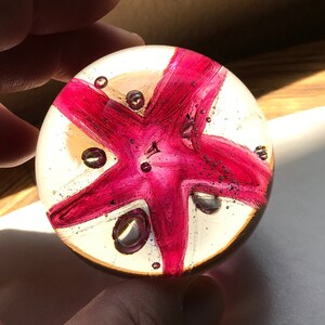 May include: A clear glass paperweight with a pink starfish design inside. The starfish has five points and is surrounded by small air bubbles.