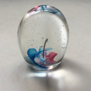 Può includere: Un fermacarte di vetro trasparente con vortici blu e rosa all'interno. Il fermacarte è pieno di piccole bolle d'aria.