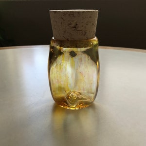 Puede incluir: Un tarro de vidrio transparente con tapa de corcho. El tarro tiene un acabado iridiscente amarillo y naranja. El tarro está sobre una superficie blanca.