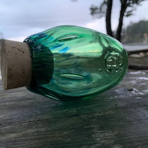 May include: A green glass bottle with a cork stopper. The bottle has a rounded shape and is filled with a swirling pattern of green and blue colors. The cork is a light brown color. The bottle is resting on a wooden surface.