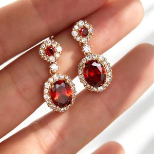 Può includere: Un paio di orecchini pendenti dorati con pietre preziose rosse ovali, circondate da piccoli cristalli trasparenti. Gli orecchini hanno un design floreale nella parte superiore e un alone simile attorno alla pietra principale. Le pietre misurano circa 1,5 cm.