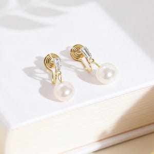 Puede incluir: Un par de pendientes dorados de clip con una perla colgante y un acento rectangular transparente. Los pendientes descansan sobre una superficie blanca, con una sombra de hojas en el fondo. Joyería clásica.