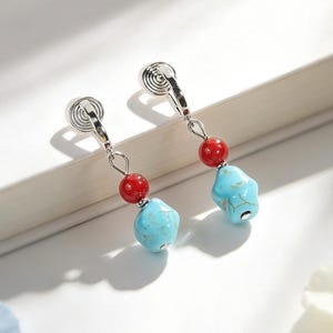 May include: A pair of silver-toned clip-on earrings featuring a small, round, glossy red bead suspended above a larger, irregular, light blue turquoise-colored stone bead with natural veining patterns, attached to a circular silver-toned clip mechanism.
