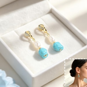 May include: A pair of gold-tone clip-on earrings featuring a white freshwater pearl and a small, irregular, bright turquoise stone. The earrings are displayed inside a white jewelry box, with an inset showing the earrings worn on an earlobe.