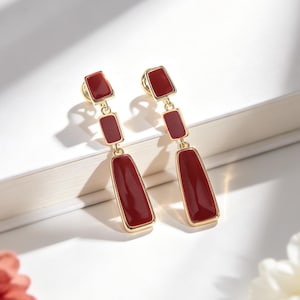 May include: A pair of gold-tone drop earrings featuring three connected rectangular segments with smooth, deep red enamel inlays. The earrings have a polished metallic finish and a vertical design that hangs from a small top post.