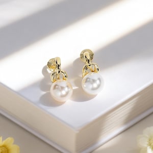 Op de afbeelding: Een paar goudkleurige oorbellen met een pareldruppelontwerp. De oorbellen hebben een decoratieve bovenkant met een kleine lus en een grote, ronde, witte parel. Gepresenteerd op een wit oppervlak met zachte verlichting.