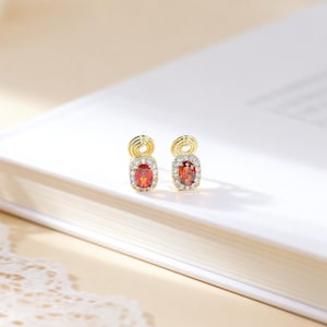 以下が含まれることがあります： 長方形のオレンジがかった赤色の宝石を中央に、小さな透明な石で囲まれた、金のイヤリングのペア。 イヤリングの上部には装飾的な金のループが付いています。 ジュエリーは白い表面に表示されています。