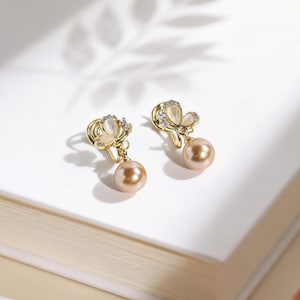 Puede incluir: Un par de pendientes de clip dorados con una parte superior en forma de mariposa adornada con pequeñas piedras transparentes y una perla redonda de color champán colgante. Los pendientes tienen un acabado de metal pulido.