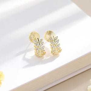 May include: A pair of gold-colored earrings with a leaf design, encrusted with small, clear gemstones. The earrings have a post-back closure and are set against a white background.