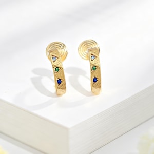 May include: A pair of gold hoop earrings featuring a textured surface and three small triangular inlays. Each earring is set with a light blue, a green, and a dark blue gemstone arranged vertically along the front of the curved metal band.