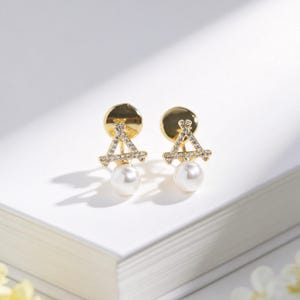 May include: A pair of gold-toned earrings with a geometric design. Each earring features a small, round stud, a triangle shape adorned with small crystals, and a dangling white pearl. The earrings are displayed on a white surface.