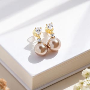 Könnte beinhalten: Ein Paar goldfarbene Ohrhänger mit einem birnenförmigen, klaren Stein oben und einem kleinen quadratischen, klaren Stein darunter. Die Ohrringe haben große, runde, hellbraune Perlen. Die Ohrringe werden auf einer weißen Oberfläche präsentiert.