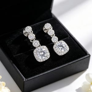 May include: A pair of silver-toned drop earrings featuring a square-cut central stone surrounded by a halo of smaller round stones. Each earring has two additional circular links set with small stones leading to the post back closure.