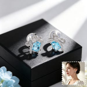 May include: A pair of silver-toned stud earrings featuring a central rectangular light blue gemstone suspended from a bow-shaped setting encrusted with small clear stones. The earrings include clear plastic backings and are displayed on a black box.