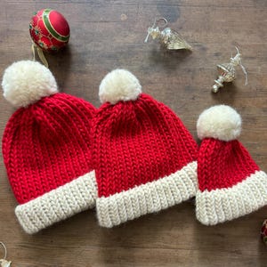 May include: Three red knit hats with thick cream-colored ribbed brims and matching cream-colored pom-poms on top. The hats are arranged in a row, featuring a chunky knit texture. Each hat is topped with a soft, round, cream-colored pom-pom.