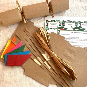 May include: A craft kit for making six Christmas crackers. The kit includes brown paper tubes, brown paper strips, brown paper diamond shapes, colorful paper flags, and brown raffia ribbon. The instructions on the card read "The Dorothy Days - Cracker Kit".