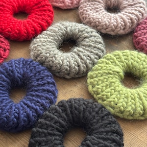May include: A collection of handmade, chunky knit scrunchies in various colors, including red, pink, gray, blue, green, and black. These hair accessories are arranged on a wooden surface, showcasing their textured, circular design.