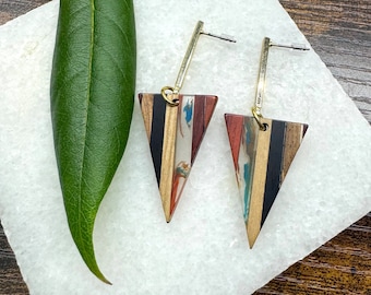Könnte beinhalten: Ein Paar dreieckige Ohrringe aus Holz mit einem goldfarbenen Metallstift. Die Ohrringe haben ein mehrfarbiges Holzmuster mit schwarzen Streifen.