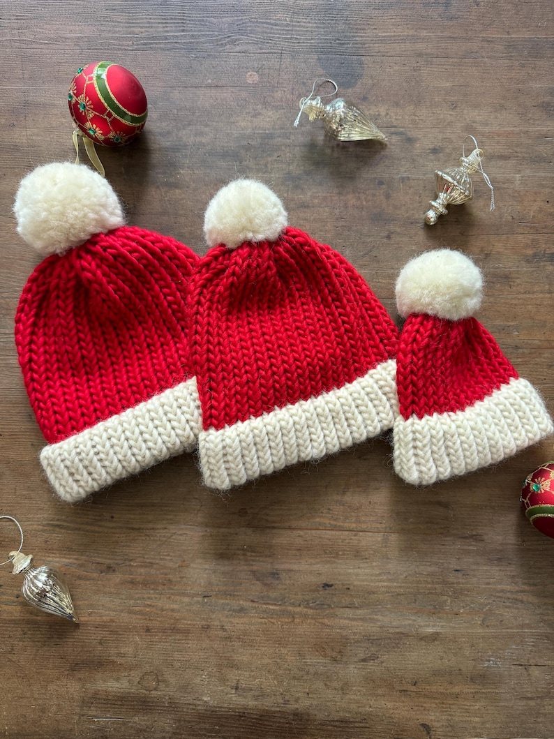May include: Three red knit hats with thick cream-colored ribbed brims and matching cream-colored pom-poms on top. The hats are arranged in a row, featuring a chunky knit texture. Each hat is topped with a soft, round, cream-colored pom-pom.