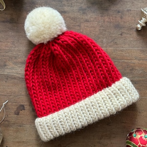 May include: A bright red knit beanie featuring a thick, cream-colored ribbed cuff at the base and a large, fluffy cream-colored pom-pom attached to the top. The hat is constructed with a chunky yarn texture and a classic vertical knit pattern.