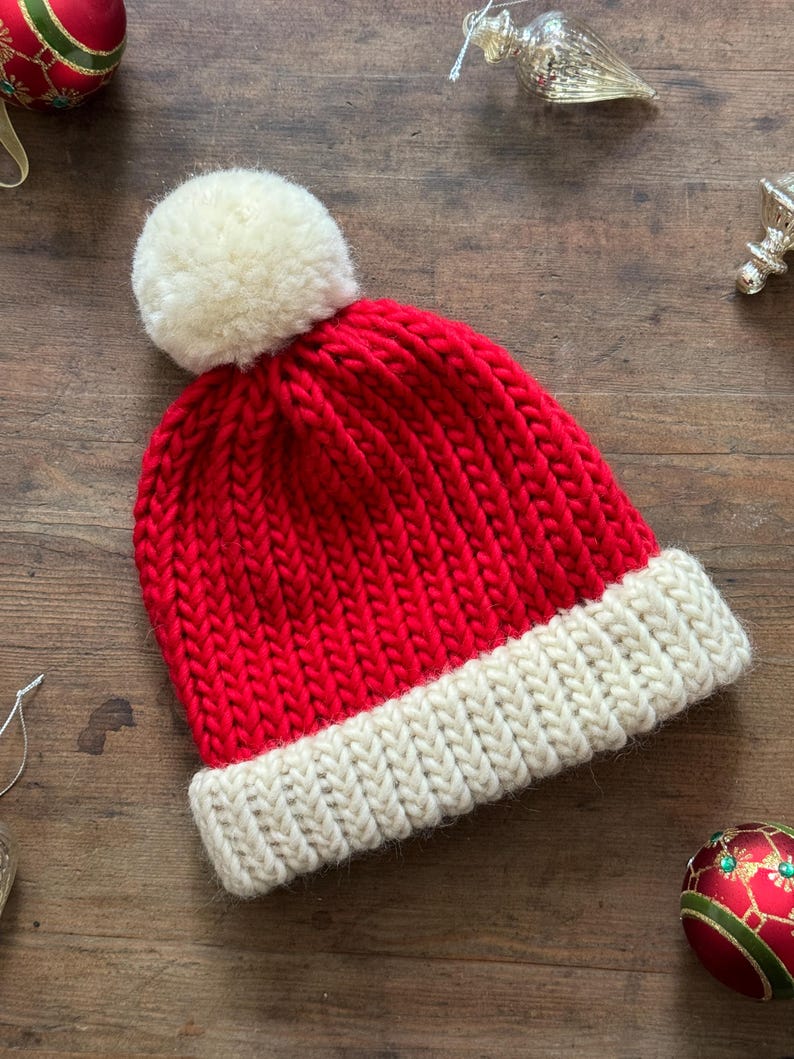 May include: A bright red knit beanie featuring a thick, cream-colored ribbed cuff at the base and a large, fluffy cream-colored pom-pom attached to the top. The hat is constructed with a chunky yarn texture and a classic vertical knit pattern.