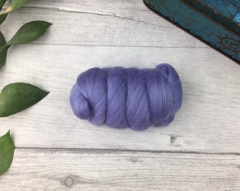 Puede incluir: Un fardo de lana de color lavanda suave, ideal para proyectos de arte textil. La lana está dispuesta en una forma cilíndrica compacta, con una superficie ligeramente texturizada. El fondo es una superficie de madera de color claro.