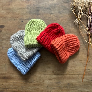 Op de afbeelding: Een verzameling van vijf gebreide mutsen in verschillende kleuren: lichtblauw, grijs, groen, rood en oranje. De mutsen zijn gemaakt van een dikke brei en liggen op een houten ondergrond. Een winteraccessoire.
