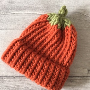 Puede incluir: Un gorro de punto de color naranja brillante con una parte superior de punto verde que se asemeja a una calabaza.