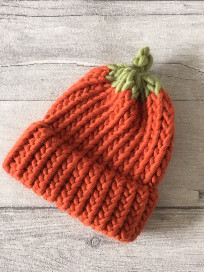Puede incluir: Un gorro de punto de color naranja brillante con una parte superior de punto verde que se asemeja a una calabaza.