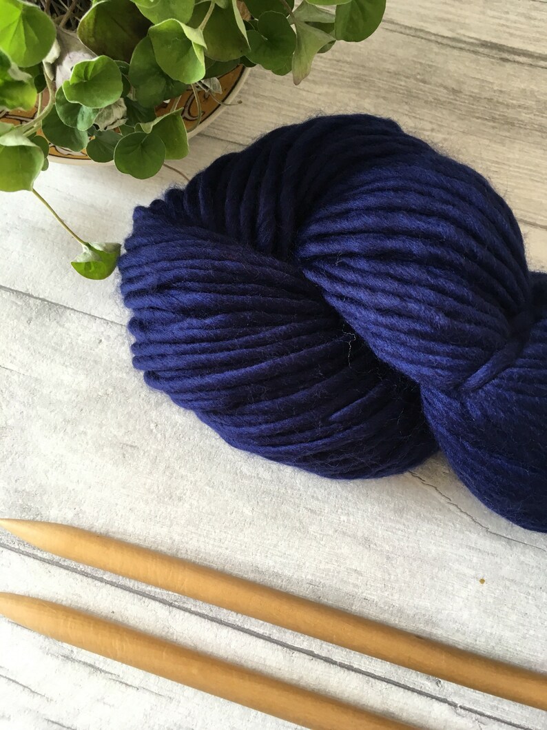 Puede incluir: Un ovillo de lana azul oscuro con dos agujas de tejer de madera.