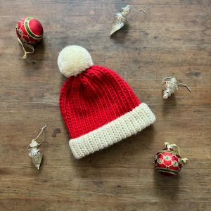 May include: A bright red knit beanie with a thick, cream-colored ribbed cuff and a matching cream-colored pom-pom on top. The hat is surrounded by two round red and green patterned ornaments and two silver-toned teardrop-shaped ornaments.
