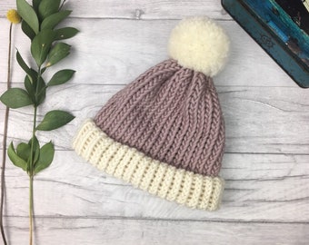 Puede incluir: Un gorro de invierno tejido con una corona lila claro y un borde color crema. El gorro tiene un gran pompón color crema en la parte superior. Hecho de hilo grueso y texturizado, perfecto para los días fríos. Medidas en centímetros.